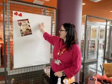 BUGlentynki w Bibliotece Głównej - II edycja. Na zdjęciu organizatorka objaśnia przy białej tablicy z plakatem przewodnim, że na niej można wpisać, za co kochamy bibliotekę. 